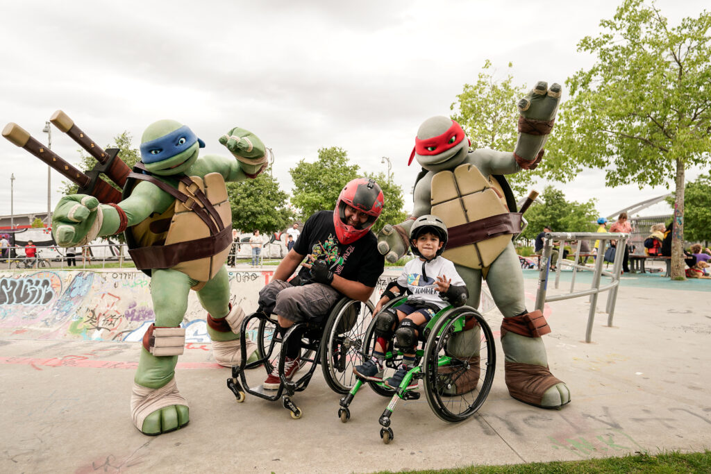 Ein Mann im Rollstuhl und ein Kind im Rollstuhl stehen im Skatepark zwischen zwei Teenage Mutant Ninja Turtles in Lebensgröße.