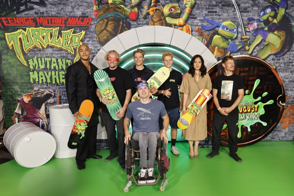 Sechs Männer, davon einer im Rollstuhl und eine Frau stehen vor einer Wand im Ninja Turtles Design.