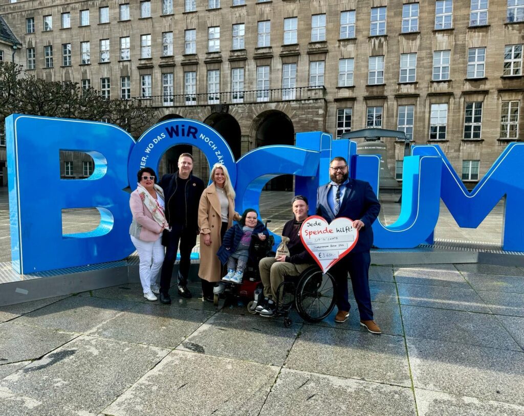 Fünf Menschen, davon zwei im Rollstuhl stehen vor großen Lettern zusammen. Jemand hält ein Herz auf dem Deine Spende hilft steht. Die großen Buchstaben im Hintergrund formen das Wort Bochum.