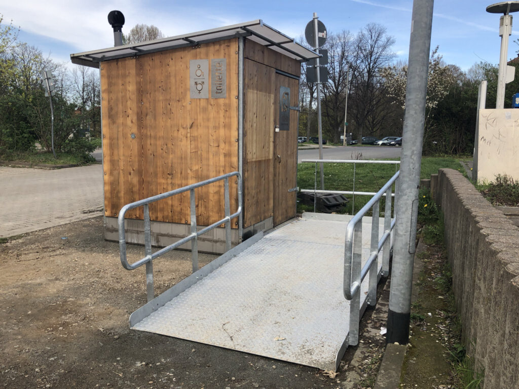 Ein hölzernes Toilettenhäuschen mit Rollstuhlrampe.