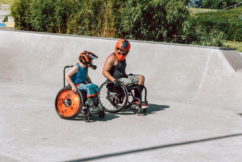 Ein erwachsener Rollstuhlfahrer neben einem Jungen im Rollstuhl im Skatepark. Beide haben Helme auf. 