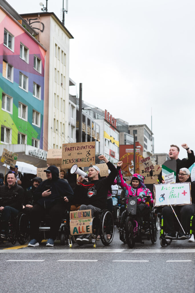 Eine Gruppe von Menschen, davon viele mit Rollstühlen demonstriert mit Schildern auf der Straße. Sie heben die Faust in die Luft. Ein Rollstuhlfahrer spricht durch ein Megaphon.
