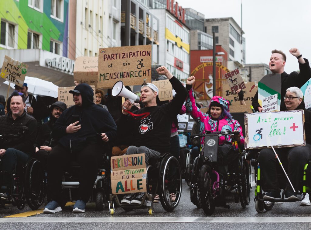Eine Gruppe von Menschen, davon viele mit Rollstühlen demonstriert mit Schildern auf der Straße. Sie heben die Faust in die Luft. Ein Rollstuhlfahrer spricht durch ein Megaphon.