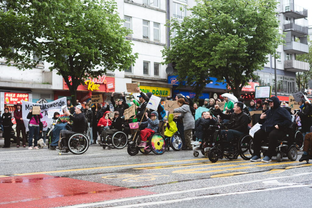 Eine Gruppe von Menschen, davon viele mit Rollstühlen demonstriert mit Schildern auf der Straße. 