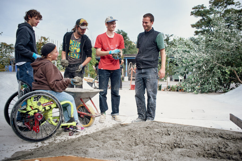 Vier Männer und eine Rollstuhlfahrerin stehen auf einer Baustelle mit Zement in der Hand.