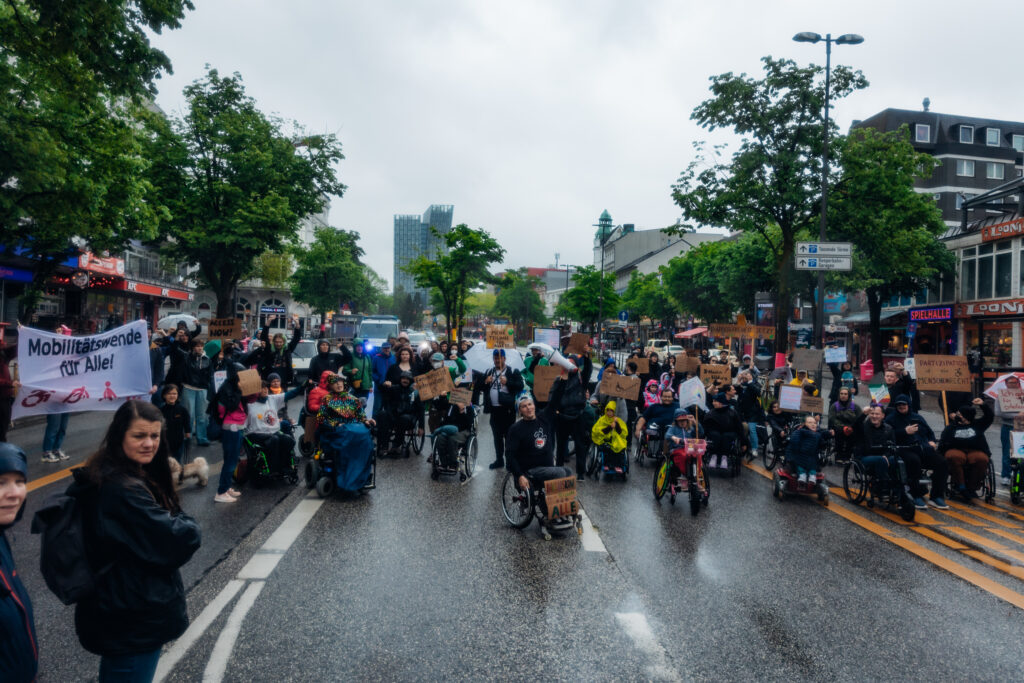 Eine Gruppe von Menschen, davon viele mit Rollstühlen demonstriert mit Schildern auf der Straße. 