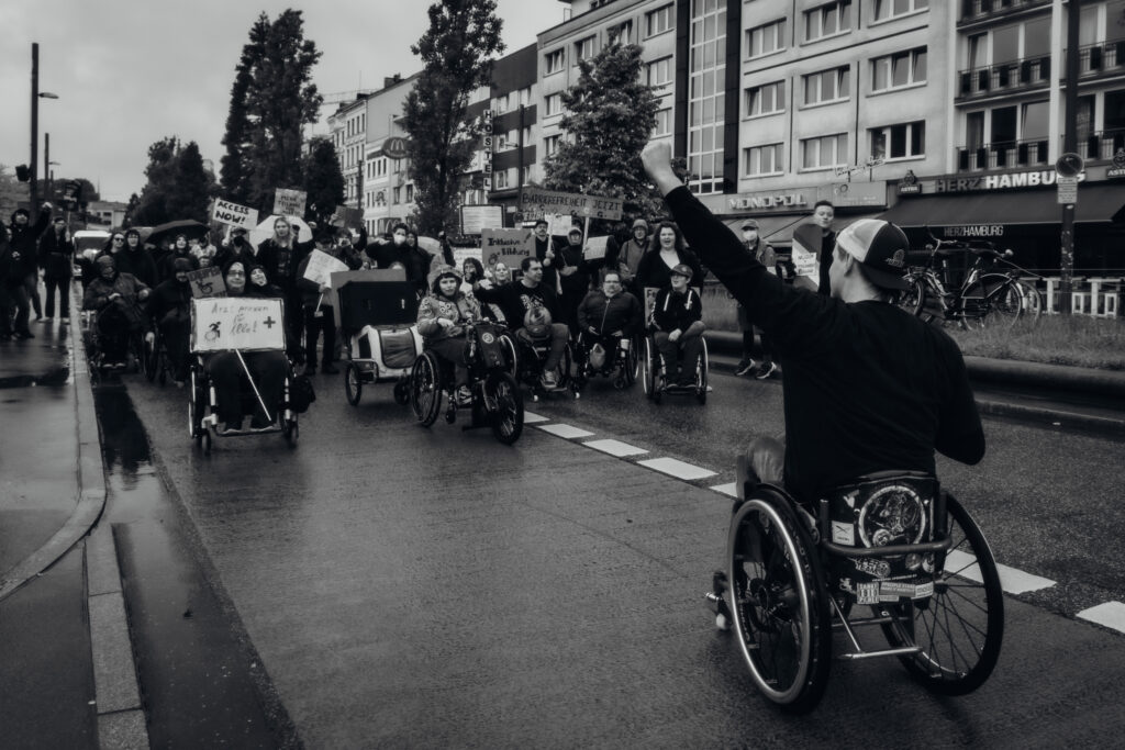 Eine Gruppe von Menschen, davon viele mit Rollstühlen demonstriert mit Schildern auf der Straße. Ein Schwarz Weiß Foto. Ein Rollstuhlfahrer steht vor der Gruppe und streckt die Faust in die Luft.