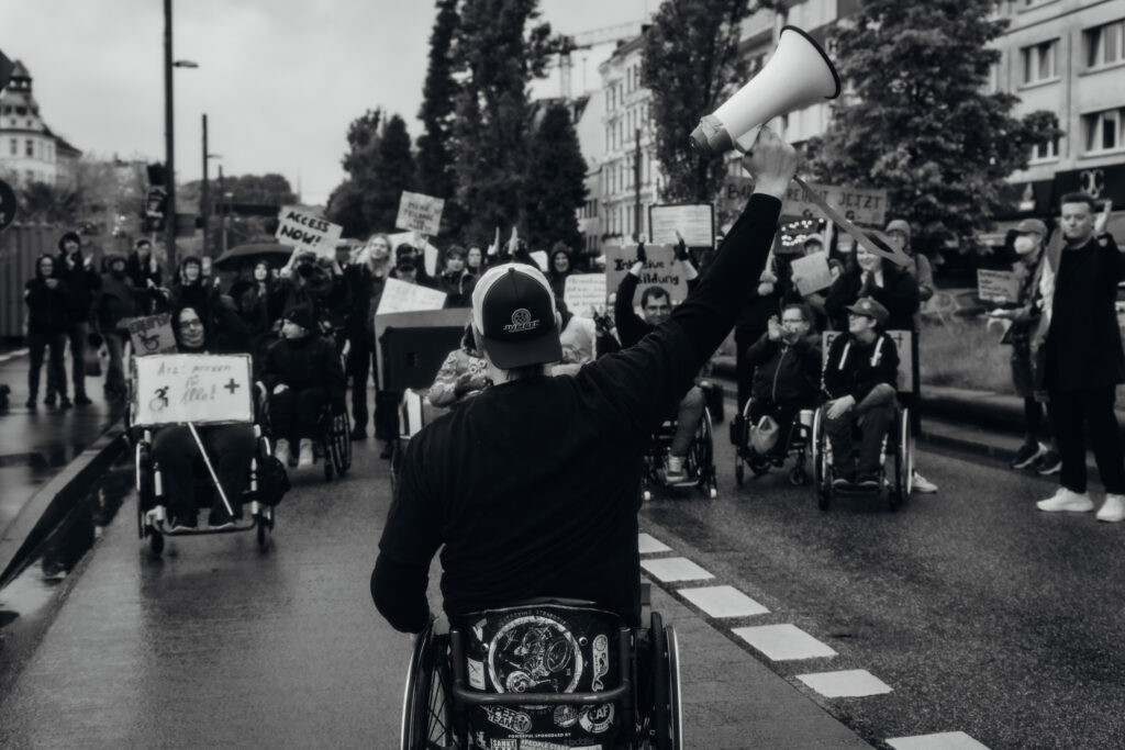 Eine Gruppe von Menschen, davon viele mit Rollstühlen demonstriert mit Schildern auf der Straße. Ein Schwarz Weiß Foto. Ein Rollstuhlfahrer steht vor der Gruppe und streckt ein Megaphon in die Luft.