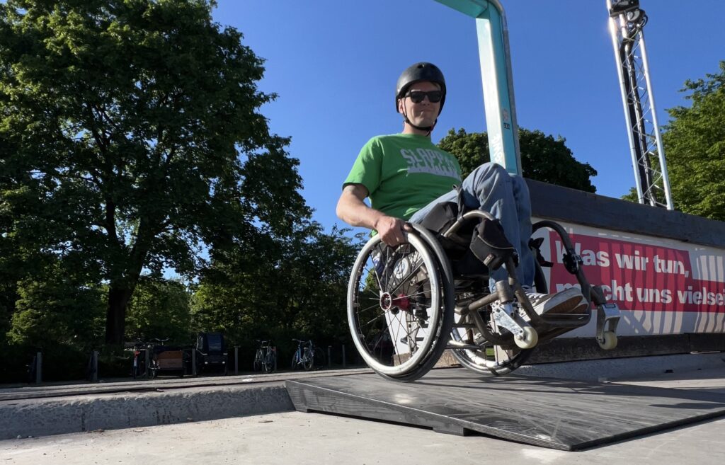 Ein Rollstuhlfahrer steht auf einer kleinen Rampe, die eine Bordsteinkante überbrückt.