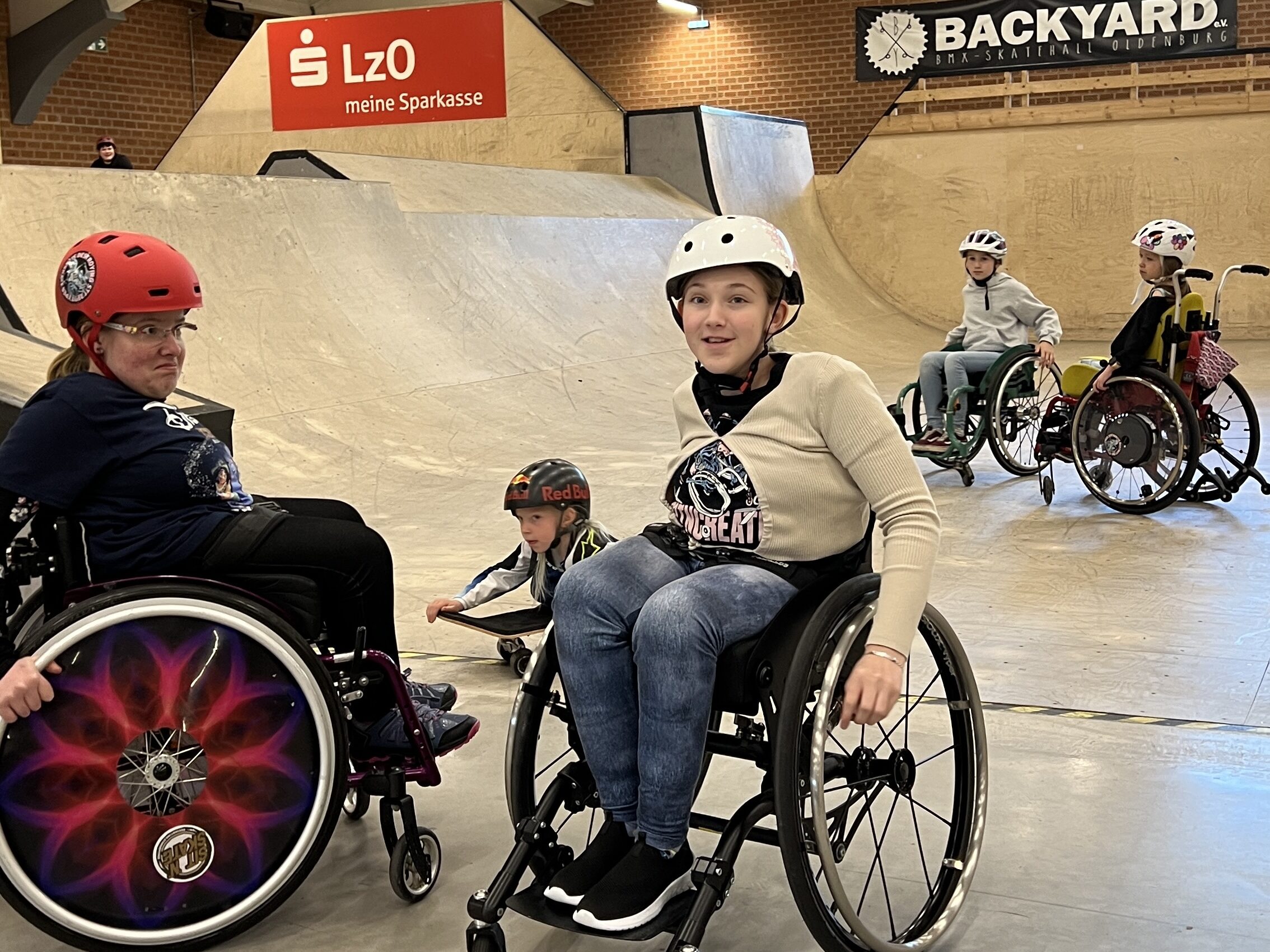 Kinder im Rollstuhl, sowie sitzend auf einem Skateboard in einer Skatehalle. 