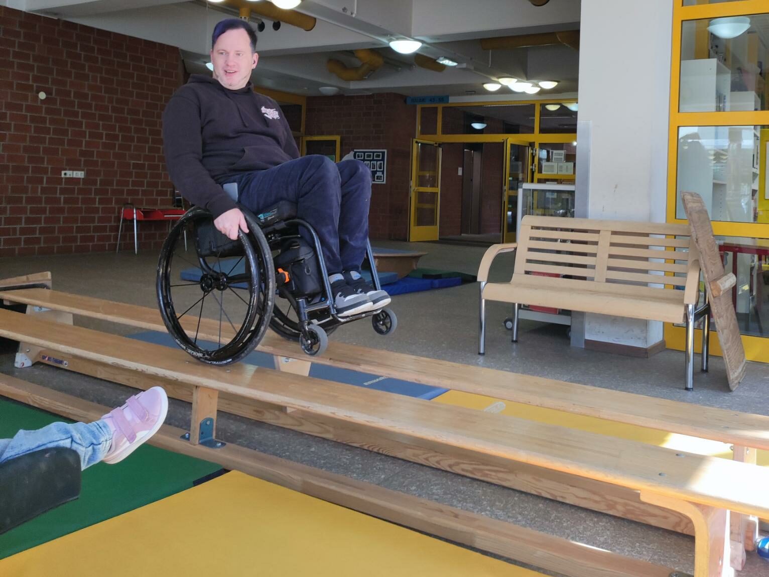 Ein Rollstuhlfahrer balanciert auf den Hinterrädern während er mit dem Rollstuhl über zwei neben einander stehende Bänke fährt. 