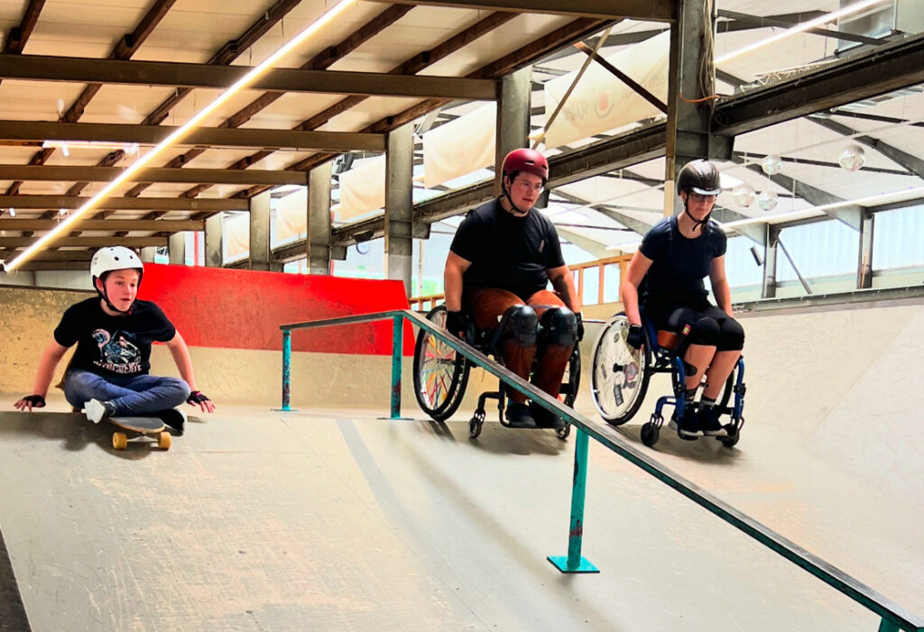 Drei Personen in Rollstühlen fahren nebeneinander über eine Rail-Rampe in einem Skatepark. Beide tragen Helme und arbeiten gemeinsam an ihren Tricks.