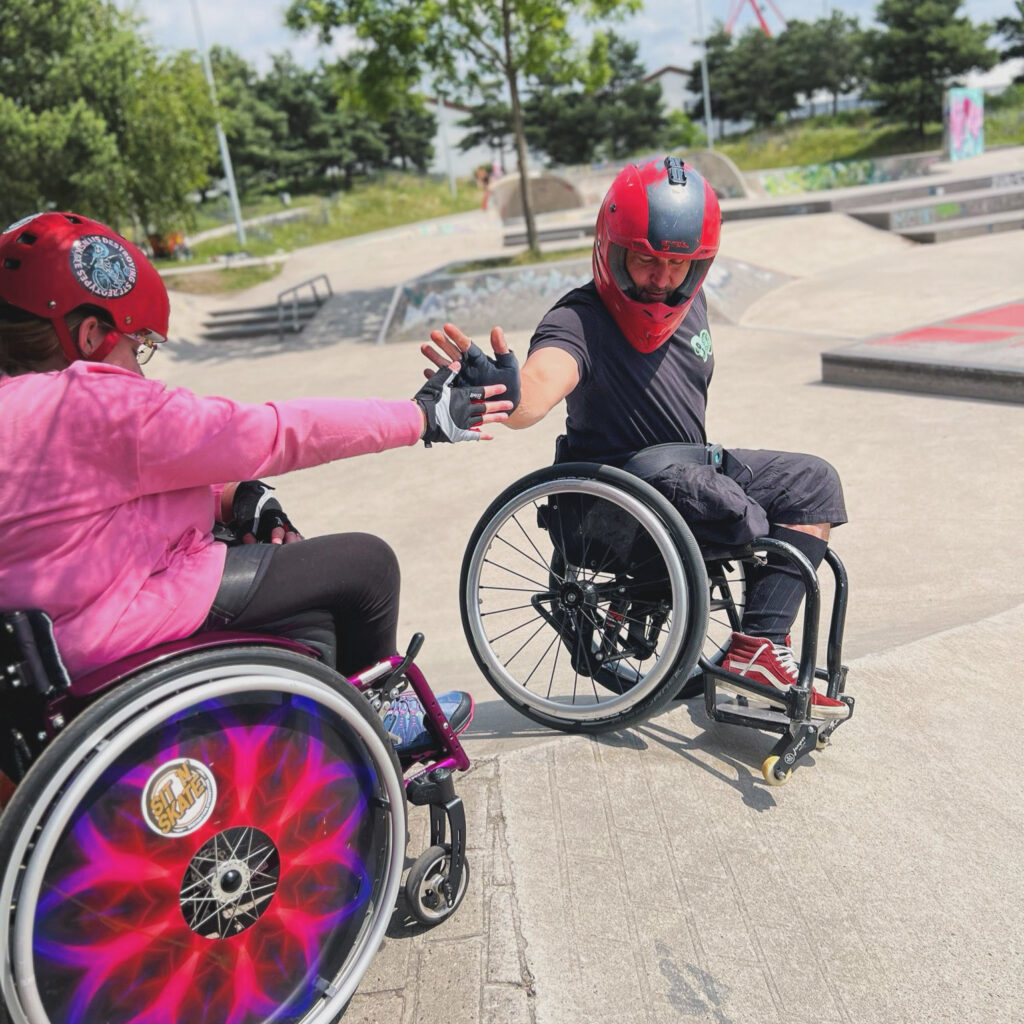 Ein Rollstuhlfahrer skatet in einem Skatepark und holt sich einen Handschlag ab von einer Rollstuhlfahrerin die oben auf der Rampe steht.