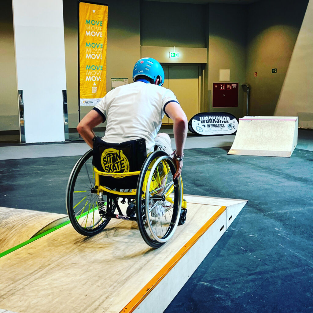 Ein Rollstuhlfahrer fährt in einer Messehalle auf einem Holzpodest, das zu einem aufgestellten Skatepark gehört.