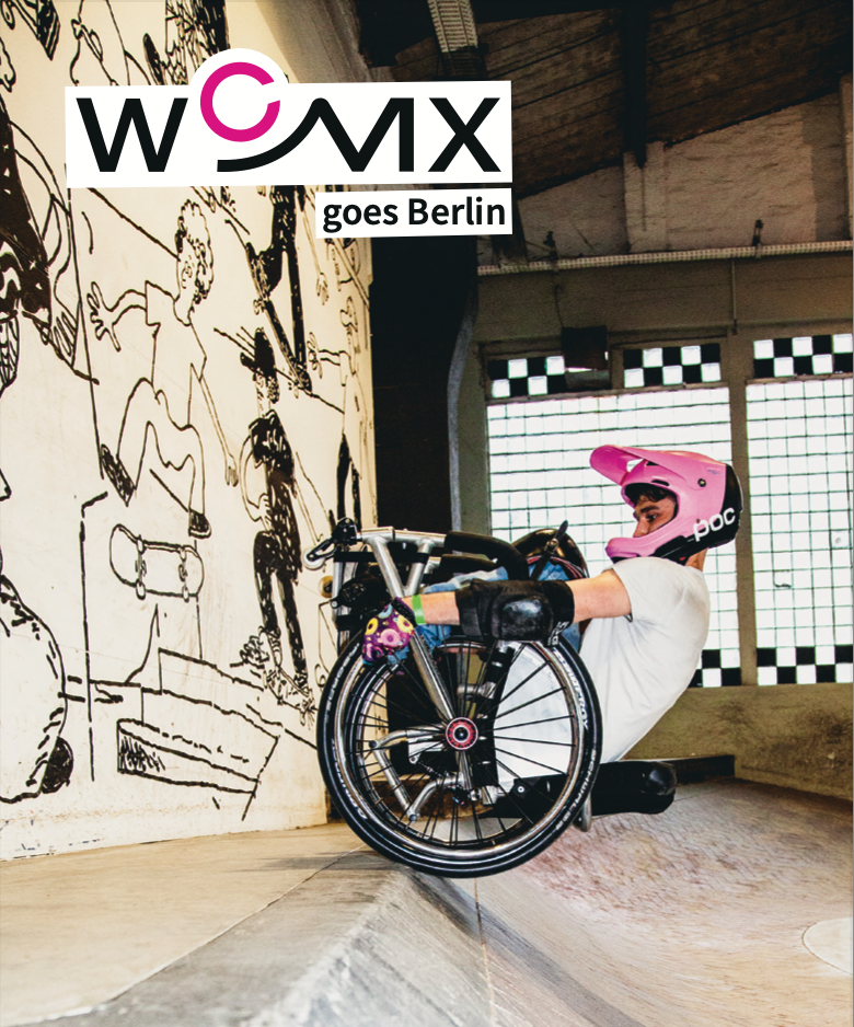 Rollstuhlfahrer macht einen Trick in einer Skatehalle und steht dabei mit den Hinterrädern an einer Kante, die Vorderräder zeigen nach oben, der Rücken über dem Boden schwebend. Oben im Bild steht WCMX goes Berlin.