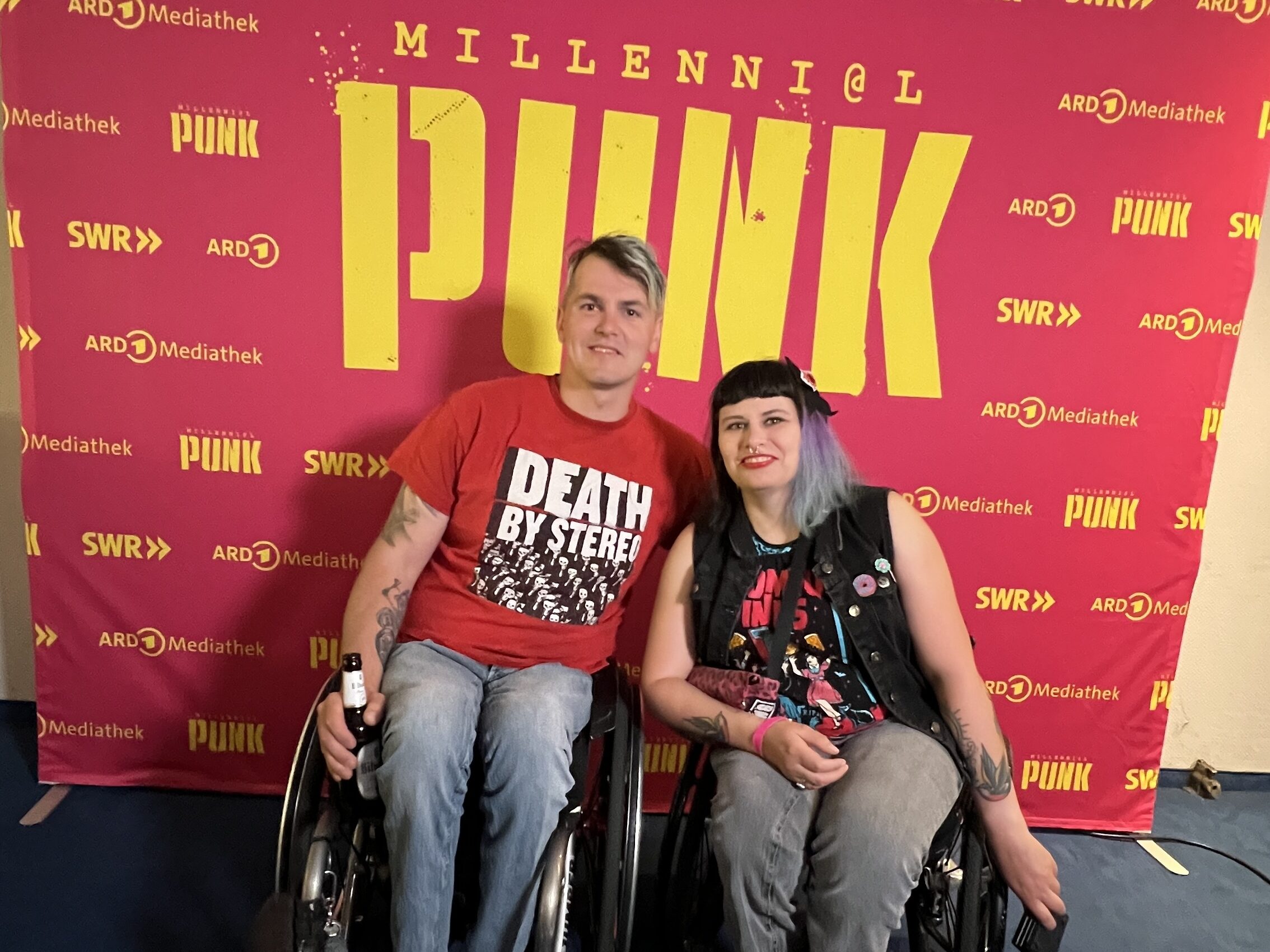 David und Lisa stehen gemeinsam vor einer pinken Fotowand auf der mit gelber Schrift Millennial Punk steht und Logos von ARD und SWR zu sehen sind.