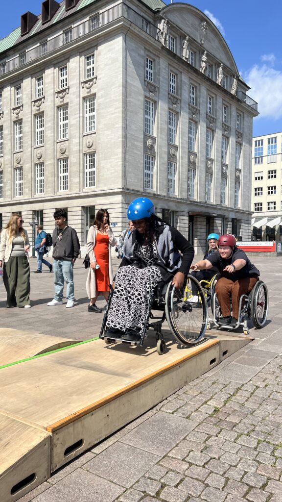 Rollstuhlfahrer hilft Schwarze Frau im Rollstuhl über eine Holzrampe, die auf einem Platz aufgestellt wurde.