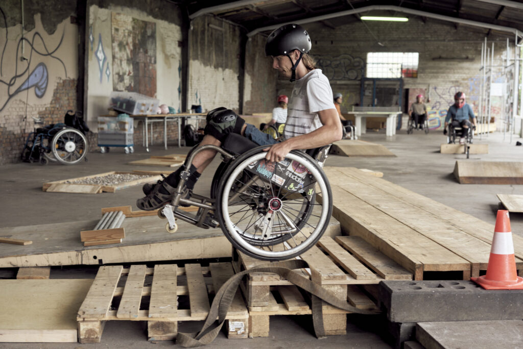 Ein Rollstuhlfahrer fährt auf den Hinterrädern balancierend ein aus Holz und Paletten gebaute Treppe herunter. Im Hintergrund weitere Rollstuhlfahrer.