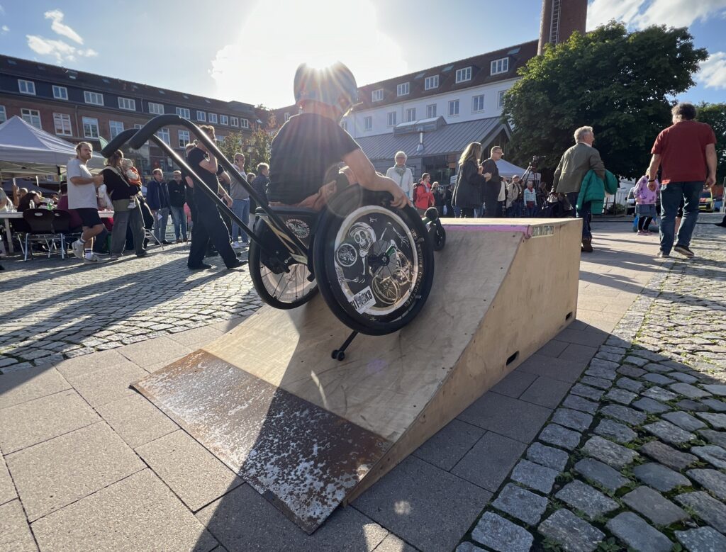 Ein Kind im Rollstuhl fährt eine auf einem Platz aufgestellte Skaterampe hinauf. Die Sonne strahlt über seinem Helm vorbei. Im Hintergrund viele Menschen. 