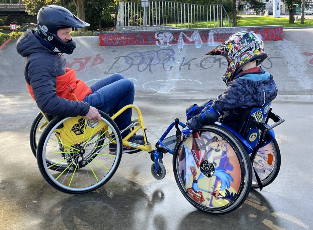 Ein erwachsener Rollstuhlfahrer und ein Kind im Rollstuhl im Skatepark. Beide balancieren auf den Hinterrädern. 