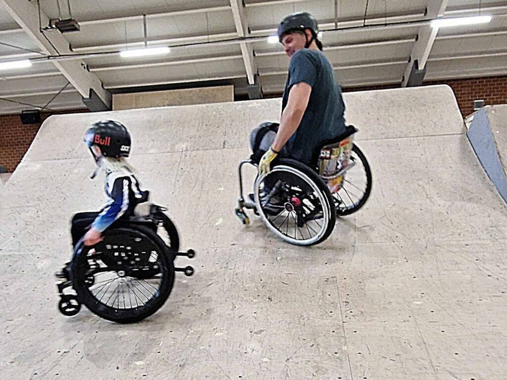 Ein Kind im Rollstuhl und ein Erwachsener, ebenfalls im Rollstuhl, stehen nebeneinander auf einer Skatepark-Rampe. Beide tragen Helme und tauschen sich aus.