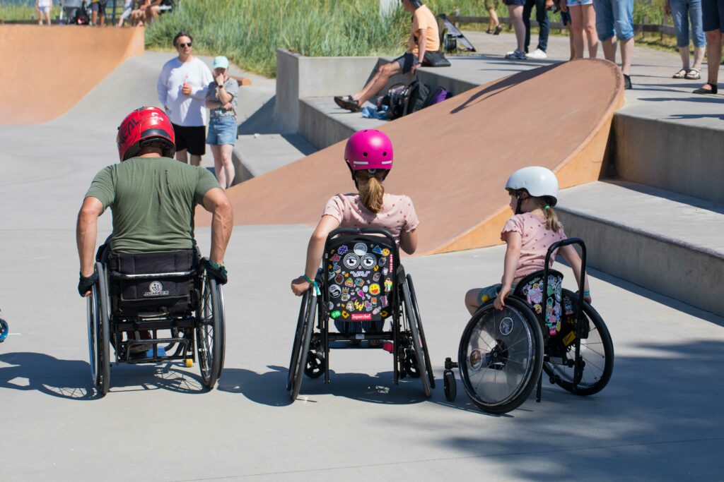 Zwei Kinder im Rollstuhl und ein erwachsener im Rollstuhl im Skatepark. 