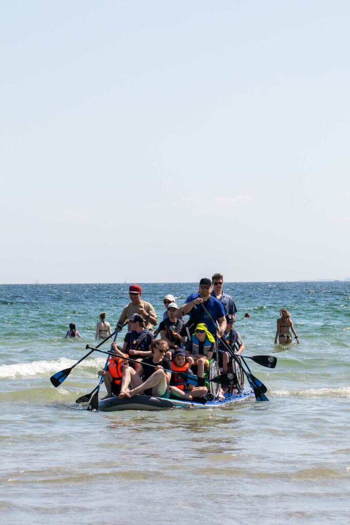 Auf einem großen SUP Board paddeln Menschen mit und ohne Rollstuhl über das Meer. 