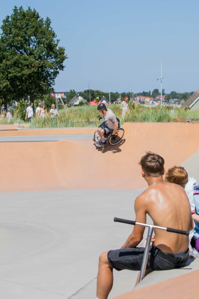 Ein Rollstuhlfahrer fährt auf einer steilen Rampe. Im Vordergrund zwei Scooter Fahrer die zuschauen. 