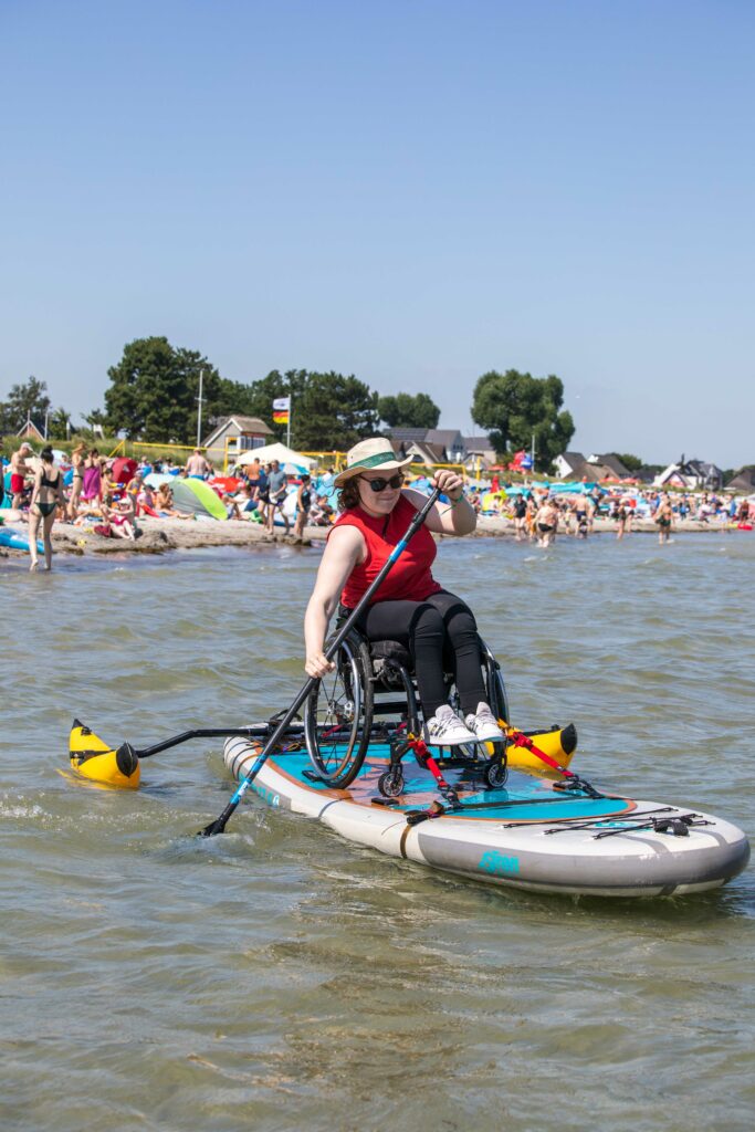 Eine Rollstuhlfahrerin, die mit ihrem Rollstuhl auf einem SUP Board über das Meer vom vollen Strand weg paddelt. Das Board hat Ausleger die wie zwei Bananen aussehen damit es nicht kippt. Der Strand ist voll mit Menschen. 