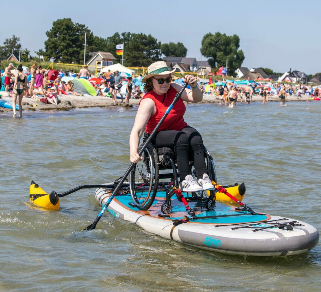 Eine Frau im Rollstuhl sitzt auf einem Stand-Up-Paddle-Board und bewegt sich mit einem Paddel auf dem Wasser. Im Hintergrund sind eine Strandkulisse und andere Menschen zu sehen.