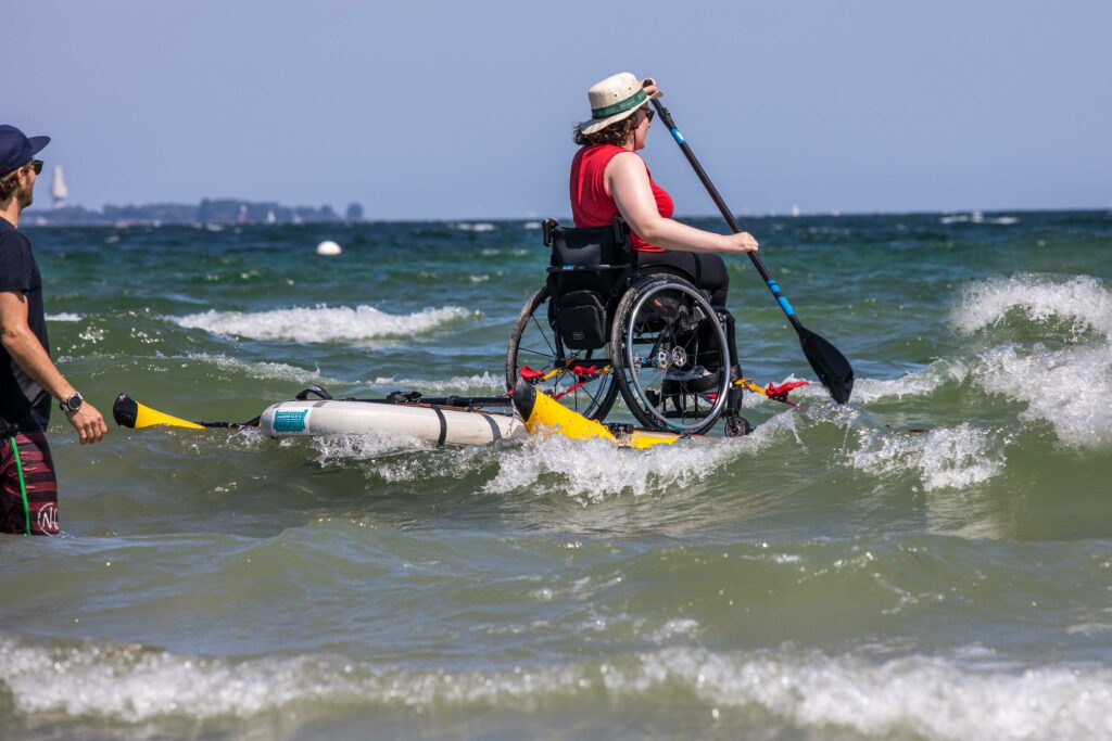 Eine Rollstuhlfahrerin, die mit ihrem Rollstuhl auf einem SUP Board über das Meer paddelt. Es sind Wellen zu sehen. 