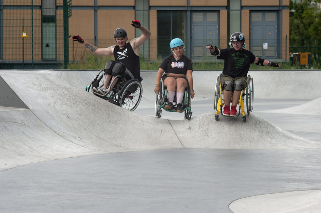 In einem Skatepark fahren drei Personen im Rollstuhl nebeneinander über ein Obstacle.