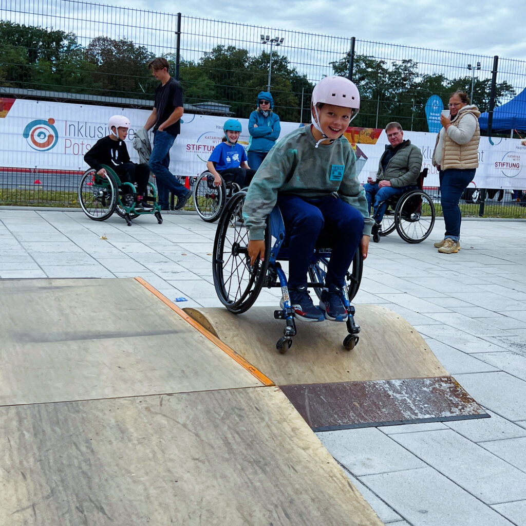 Ein Kind im Rollstuhl mit Helm fährt über ein mobiles Holzelement, dass zum Skaten auf einem Platz gestellt wurde. Im Hintergrund sieht man weitere Menschen im Rollstuhl und einen Bananen an dem Zaun, auf den die Veranstaltung Inklusiv gewinnt und das du der Katarina Witt Stiftung zu sehen sind.