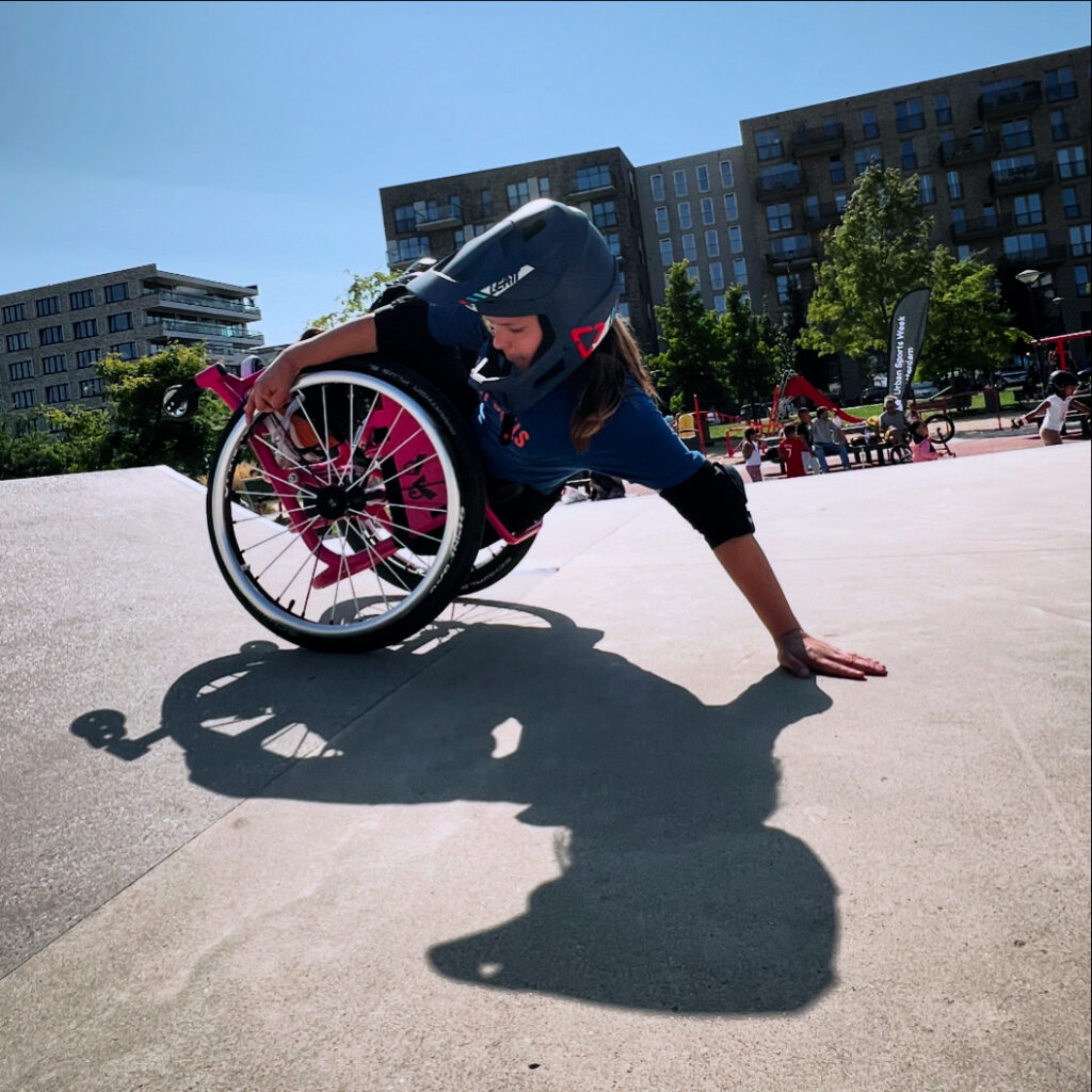 Eine Rollstuhlfahrerin im Skatepark lehnt sich weit nach hinten, sodass ihre Hand den Boden berührt. Die andere Hand am Rad. Man sieht ihren Schatten vor ihr auf dem Beton