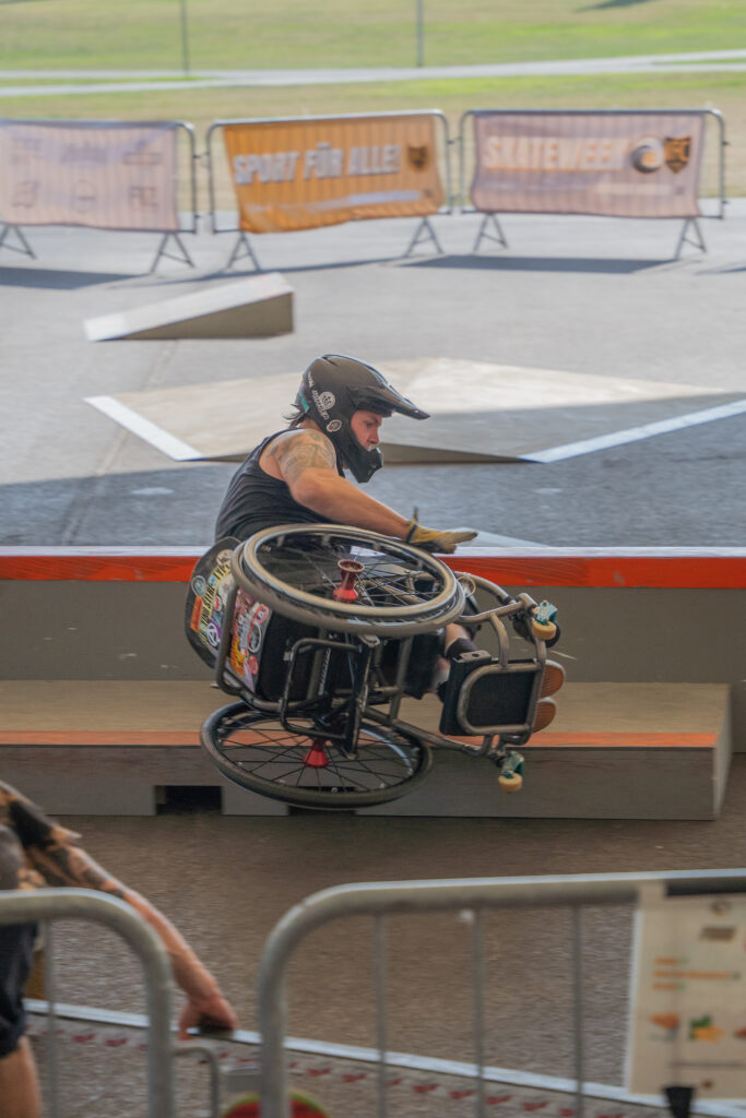 Ein Rollstuhlfahrer slidet auf der Seite mit dem Rad über ein  Curb im Skatepark. 