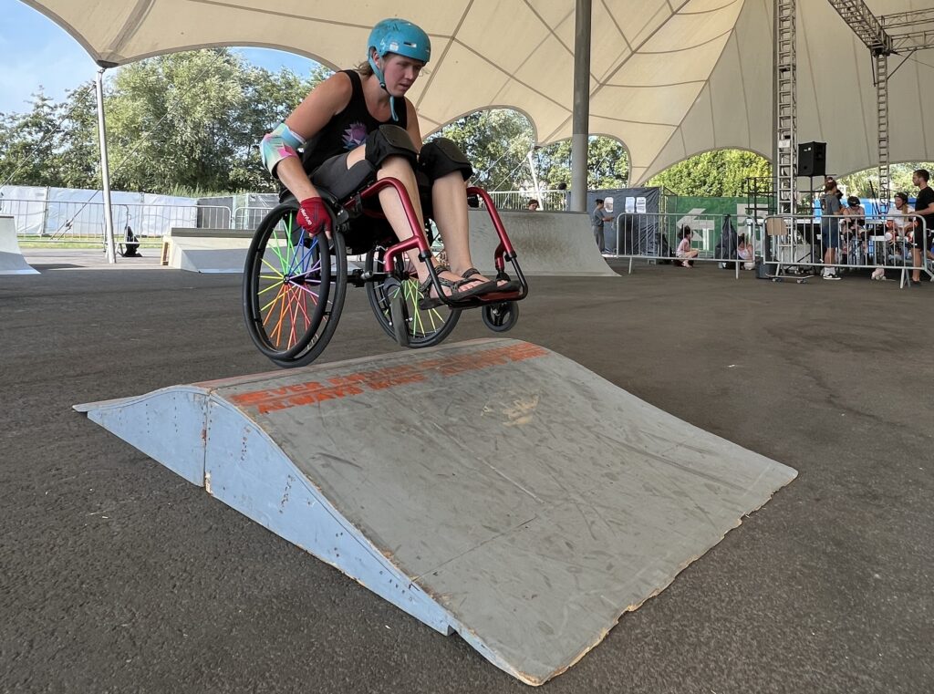 Eine Rollstuhlfahrerin fährt über eine Welle aus Holz in einem mobilen Skatepark unter einem großen Zelt.