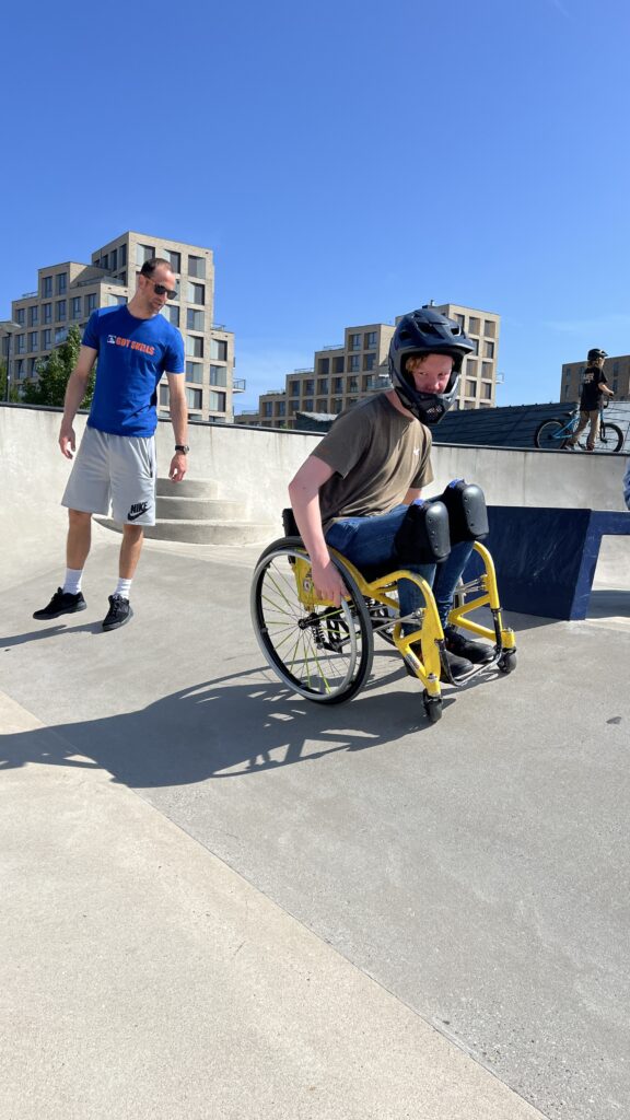 Rollstuhlfahrer im Skatepark. Dahinter steht ein Mann und beobachtet.