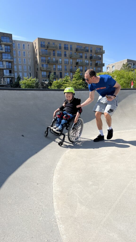 Ein Mädchen im Rollstuhl im Skatepark wird von einem Mann an einer Rampe unterstützt.