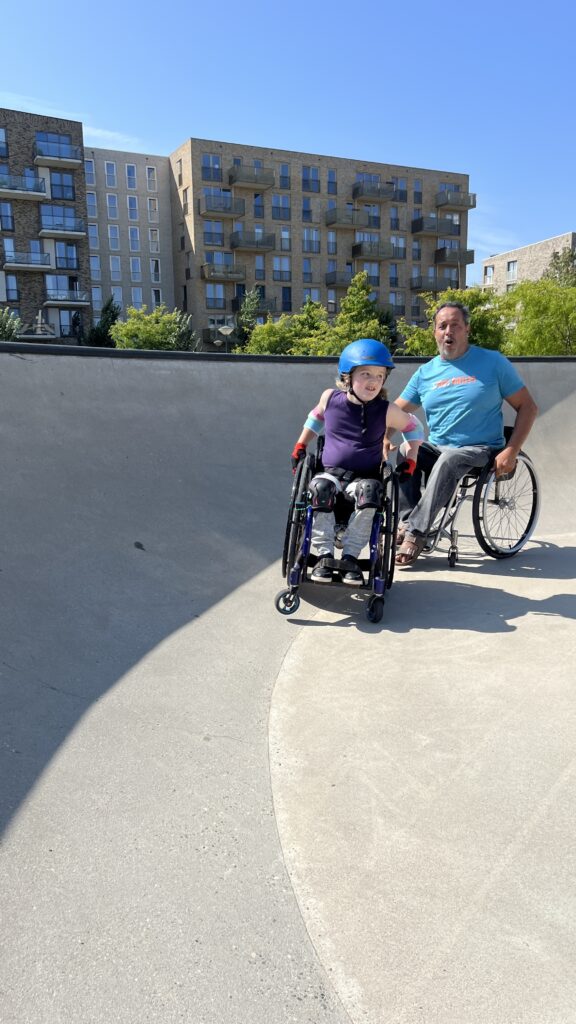 Ein Mädchen im Rollstuhl im Skatepark wird von einem Mann im Rollstuhl an einer Rampe unterstützt.