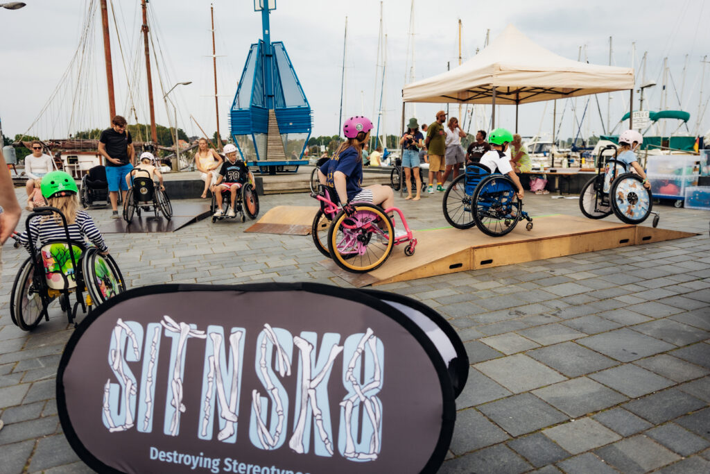 Auf einem Platz sind Pavillons und mobile Holz Skate Elemente aufgestellt, wo Rollstuhlfahrer mit Helmen rüber fahren. Vorn steht ein Banner mit den Buchstaben SIT N SKATE.