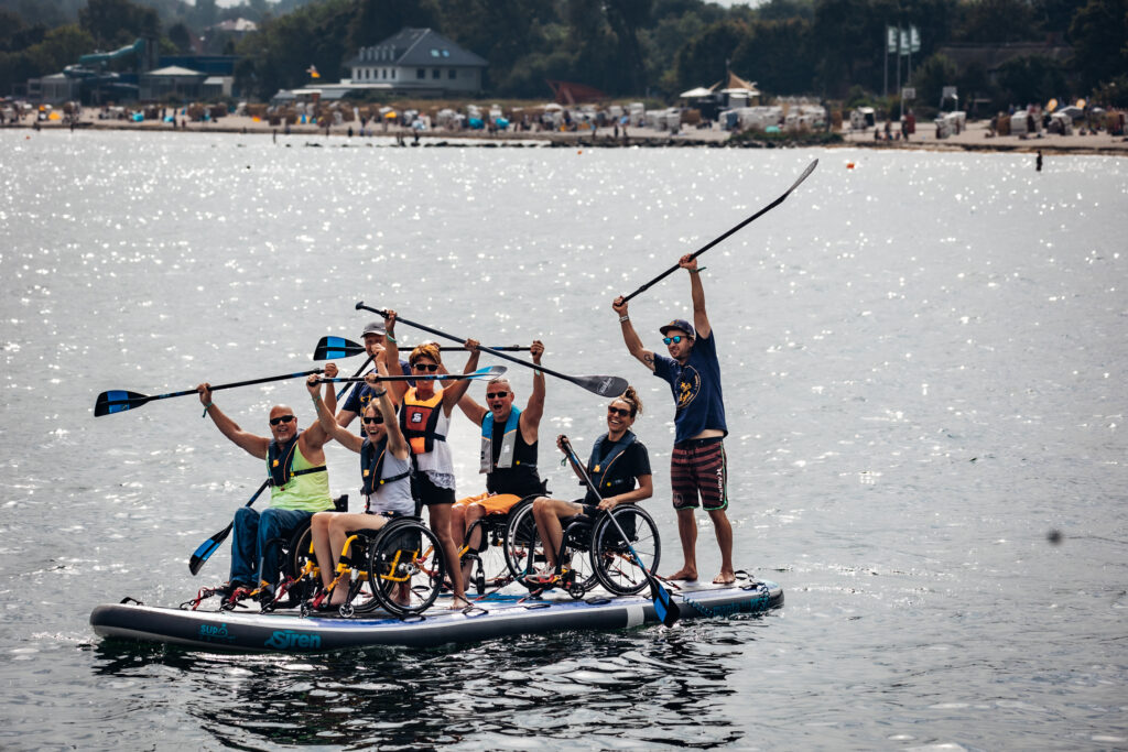 Auf einem großen SUP Board sind Menschen mit und ohne Rollstuhl auf dem Meer. Sie halten die Paddel nach oben in die Luft. Im Hintergrund sieht man den Strand mit vielen Menschen. 