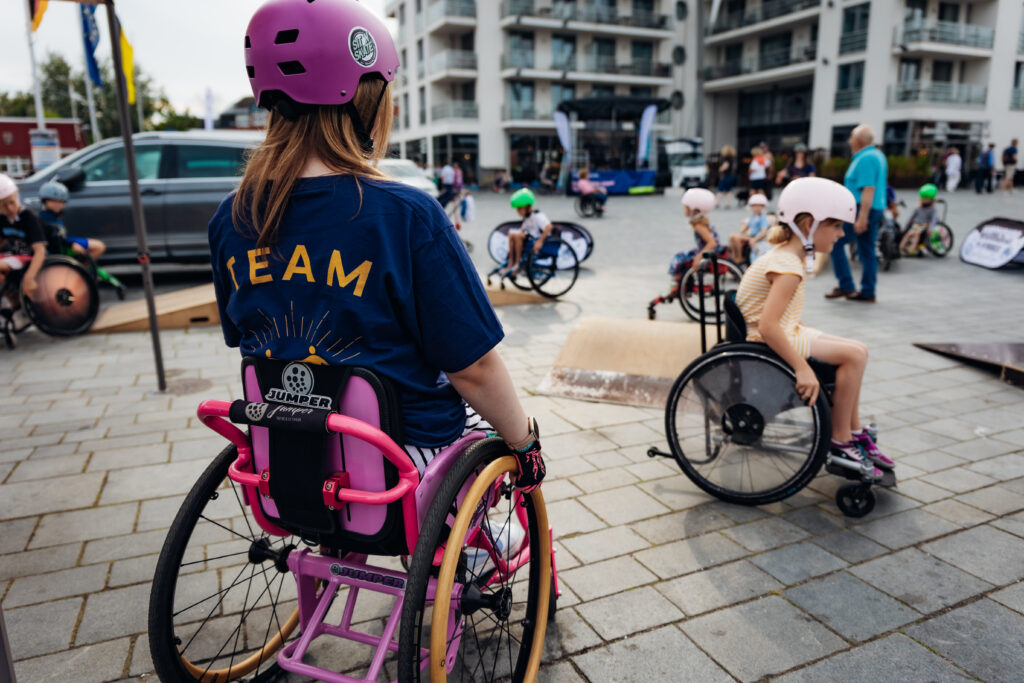 Ein Platz mit Kindern im Rollstuhl und mobilen Skate Elementen. Im Vordergrund eine Frau in einem pinken Rollstuhl. Auf ihrem Shirt auf dem Rücken steht TEAM.