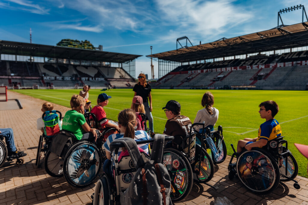 Eine Gruppe von Kindern im Rollstuhl steht im Millerntorstadion am Rasen. Eine Frau steht vor der Gruppe und erzählt etwas. Auf den Blocken im Stadion sieht man Herzen und den Spruch Kein Fußball den Faschisten.