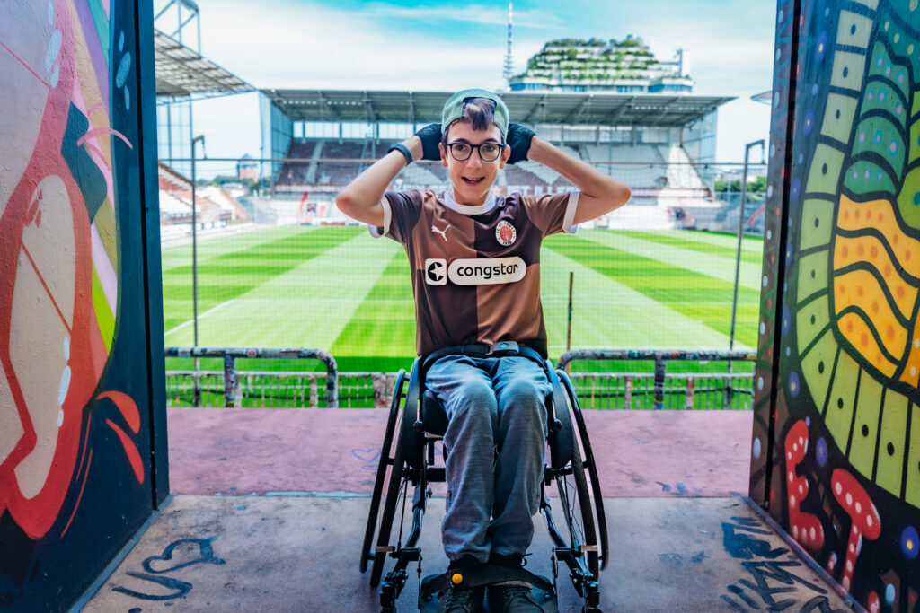 Ein Junge im Rollstuhl im St. Pauli Trikot steht auf der Tribüne im Millerntorstadion. Im Hintergrund sieht man das Spielfeld und den Hamburger Bunker mit seinem neuen grünen Dach.