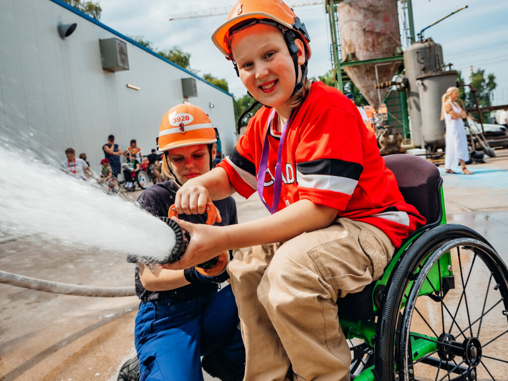 Ein Kind im Rollstuhl trägt einen orangefarbenen Helm und hält einen Feuerwehrschlauch, während eine Person im Feuerwehranzug hinter ihm den Schlauch unterstützt. Im Hintergrund sind andere Kinder und eine Feuerwehrkulisse zu sehen.