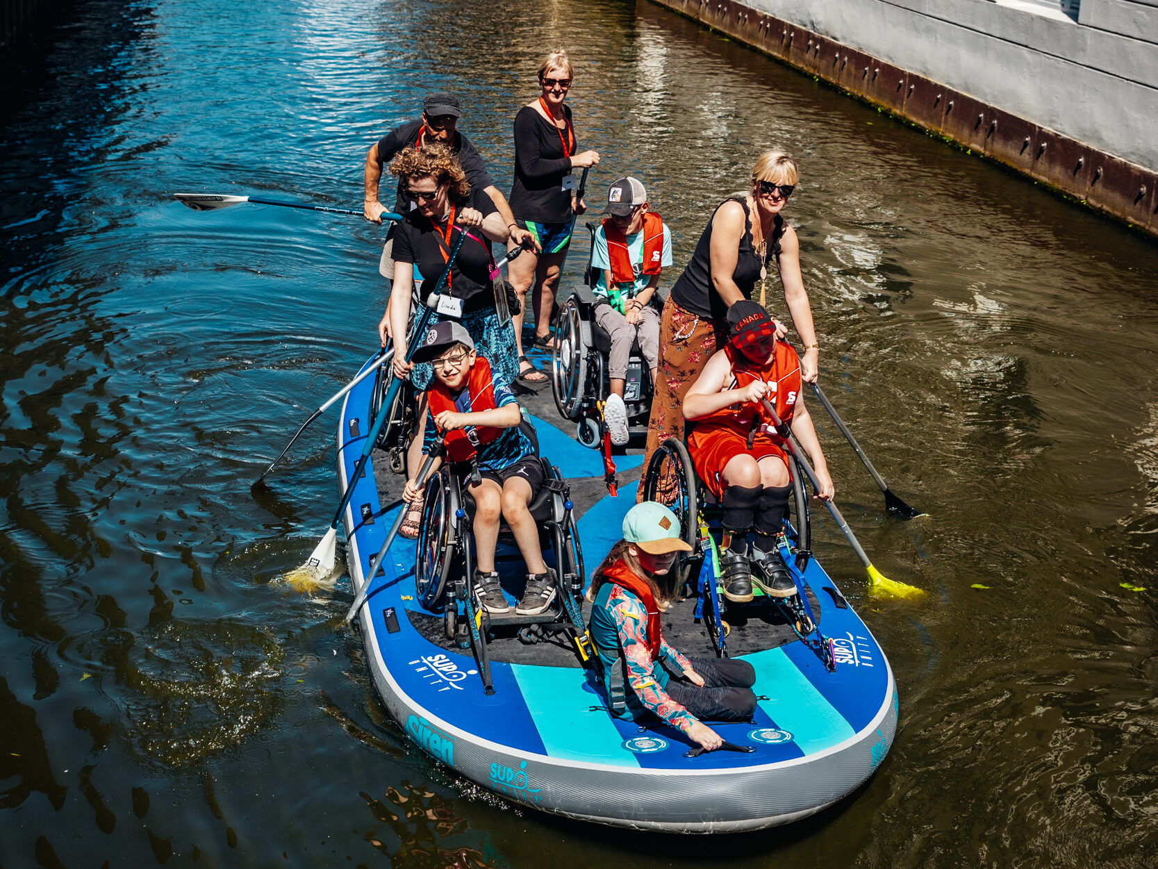 Kinder im Rollstuhl paddeln auf einem großen SUP Board auf dem Wasser. Die Rollstühle sind auf dem Board festgeschnallt. Drei erwachsene Menschen helfen mit. 