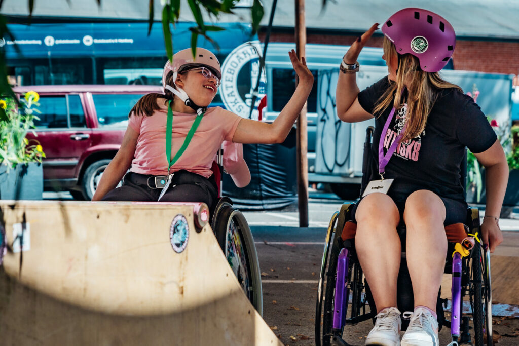 Zwei Frauen im Rollstuhl geben sich lachend ein High-Five in einem Skatepark. Beide tragen Helme, und im Hintergrund sind weitere Teilnehmende und Zuschauer zu sehen.