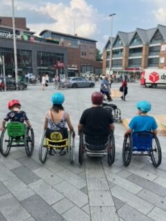 Auf einem Marktplatz stehen vier Rollstuhlfahrer mit Helmen nebeneinander. 