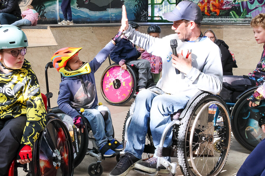 Ein Kind im Rollstuhl gibt einem Erwachsenen Rollstuhl ein HighFive in einer Skatehalle