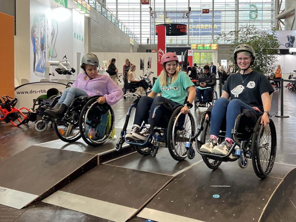 Drei Frauen im Rollstuhl stehen auf den Hinterrädern balancierend auf verschieden hohen mobilen Holzelementen in einem kleinen Skatepark in einer Messehalle. 